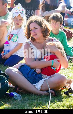 MUTTER UND KIND AUF DEM SOMMER-MUSIKFESTIVAL, REPORTAGE, GREEN MAN FESTIVAL 2013, BRECON, WALES, UK, 2013: Eine Frau in einem weißen Oberteil und Jeansshorts sitzt im Sonnenschein im Schneidersitz auf dem Rasen und hält ein kleines Kind in einem roten T-Shirt. Foto: Rob Watkins. INFO: Das Green Man Festival findet jährlich im August in den Brecon Beacons statt. Es feiert alternative Musik, Kunst, Literatur, Wissenschaft und familienfreundliche Kultur in einer wunderschönen Berglandschaft. Das Event 2013 war die 10. Jubiläumsausgabe. Stockfoto