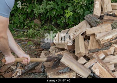 Mann hackt mit einer Axt im Hinterhof Holz. Starke Männerhände halten ein scharfes Werkzeug und schlagen auf einen Baumstamm auf einem Baumstumpf. Stapel gestapelter gehackter Feuerholzblöcke Stockfoto