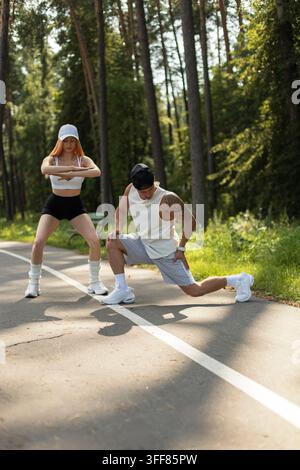 Fittes junges Paar macht Aufwärm-Dehnübungen auf einem sonnigen Parkweg im Freien. Athletischer Mann und Frau trainieren zusammen in Sportkleidung, umgeben Stockfoto