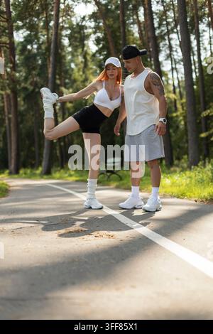 Fittes junges Paar streckt sich zusammen auf einem Parkweg im Freien. Athletische Frau balanciert auf einem Bein und dehnt die Oberschenkelmuskeln, während der unterstützende Mann dabei ist, an einem sonnigen Sommertag Stockfoto