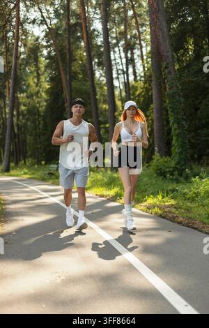Fittes junges Paar joggt Seite an Seite auf die Kamera zu auf einem gepflasterten Parkweg. Athletischer Mann und rothaarige Frau laufen in Sportkleidung während eines sonnigen Sommers Stockfoto