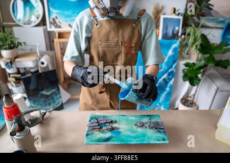 Nahaufnahme einer Künstlerin, die blaue Epoxidharz auf ein handgefertigtes abstraktes Gemälde zu Hause gießt. Stockfoto