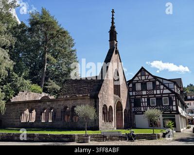 Steinkapelle von Bad Herrenalb mit hohem, verzierem Turm und gewölbtem Kreuzgang steht neben einem Fachwerkhaus, das von alten Bäumen umrahmt ist, unter einem strahlend blauen Himmel, Stockfoto