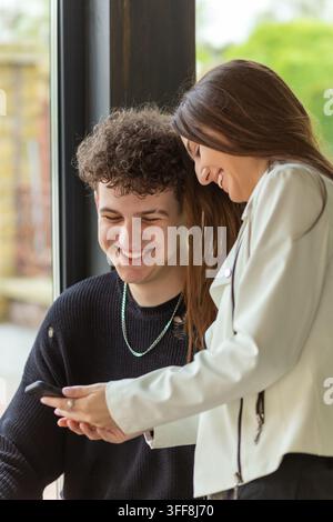 Fröhliches junges Paar lächelt am Fenster auf das Smartphone, teilt Online-Inhalte und genießt einen warmen Moment zusammen drinnen. Stockfoto