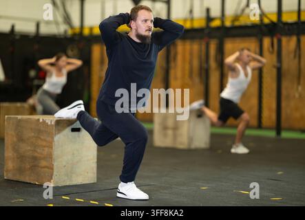 Konzentrierter junger bärtiger Mann in Sportkleidung führt bulgarische Split-Squats auf einer hölzernen Plyometrik-Box während intensivem Körpergewichts-Training im Fitnessstudio durch.. Stockfoto