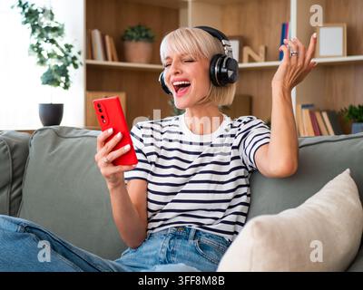 Glückliche junge Frau hört ihre Lieblingsplaylist über drahtlose Kopfhörer, während sie ihr Telefon benutzt. Stockfoto