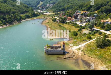 Luftbild der versunkenen Kirche St. Nikolaus, teilweise im Mavrovo-See versunken, umgeben von grünen Hügeln und Dorfhäusern in Mavrovo, Municip Stockfoto