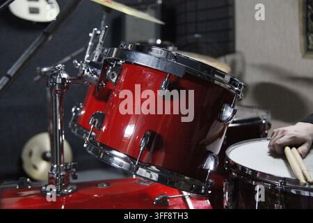 Eine Person spielt einen Beat auf einem roten Schlagzeug mit hölzernen Drumsticks. Die Szene findet in einem Musikstudio während der Abendstunden statt. Stockfoto