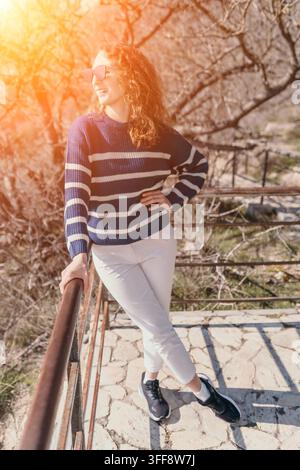 Frau lächelt im Sonnenlicht. Glückliche junge Frau mit lockigem Haar genießt einen sonnigen Tag im Freien, trägt gestreiften Pullover und Sonnenbrille Stockfoto