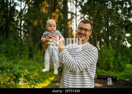 Vater hält seinen neugeborenen Sohn im Freien, schaut in die Kamera, feiert die Vaterschaft und die Familienbindung in einer Waldumgebung bei Sonnenuntergang und genießt die frische Luft Stockfoto