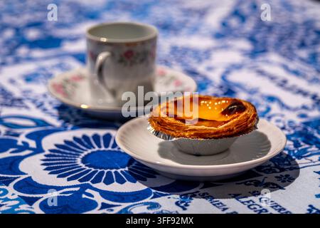 Eine pastellfarbene de nata-Krustentorte mit einer kleinen Porzellan-Kaffeetasse mit Blumenmuster auf einer blauen und weißen Tischdecke mit portugiesischen Azulejo-Motiven Stockfoto