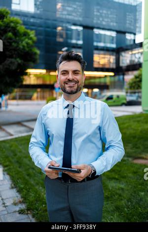 Ein Mann lächelt, während er ein Tablet vor einem modernen Gebäude hält. Er trägt ein hellblaues Hemd und eine Krawatte. Stockfoto