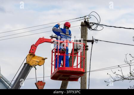 Maykop, Russland - 6. Dezember 2024: Ein Elektriker in einer blauen Latzhose von dem Energieunternehmen Rosseti und einem roten Helm installiert Stromleitungskabel o Stockfoto