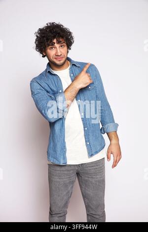 Ein sauberes mittleres Studio-Porträt eines gutaussehenden jungen Mannes mit lockigem schwarzem Haar. Er trägt ein lässiges blaues Denimhemd und lächelt wissend, während er auf Stockfoto