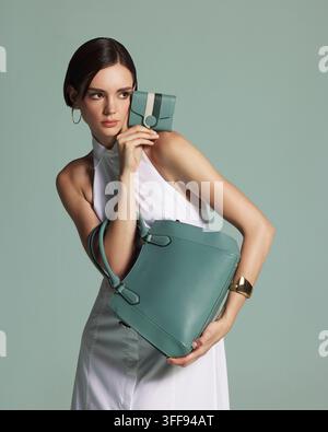 Eine schöne junge brünette Frau in einem eleganten weißen Neckholderkleid posiert mit einer hochwertigen smaragdgrünen Ledertasche auf der Schulter. Stockfoto