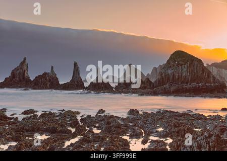 058 Lebendiger Morgen über Playa Gueirúa Beach und seiner Reihe scharfer Kliffen, die von einer dichten dunklen Wolke mit der Sonne darüber begleitet werden. Cudillero-Asturien-Spanien. Stockfoto