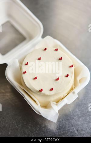 Kleine weiß gefrostete Torte verziert mit winzigen roten Herzstreuseln in einer Mitnahmeschachtel auf Metalloberfläche, mit Torte, Glasur, Weiß, Streusel-Detail, zeigt Stockfoto