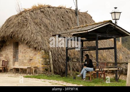 Eine junge Frau sitzt auf einem Holzstuhl unter einem kleinen rustikalen Schuppen und schaut auf ihr Handy neben einem traditionellen runden Steingebäude mit Strohdach in einem r Stockfoto