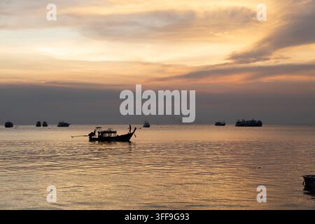 Ein silhouettiertes traditionelles Langschiff mit einer stehenden Figur ruht auf einem glitzernden goldenen Meer bei Sonnenuntergang, mit einer Flotte von festgemachten Schiffen, die den Dunst übersäen Stockfoto