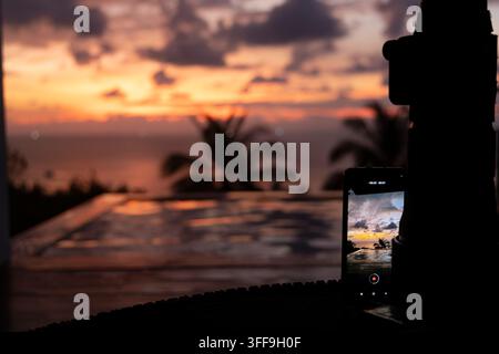 Ein Smartphone, das auf einem Stativ montiert ist, zeichnet einen lebhaften orangefarbenen und lila Sonnenuntergang über einem Infinity-Pool auf, der von silhouettierten Palmen umgeben ist, mit dem Ozean v Stockfoto