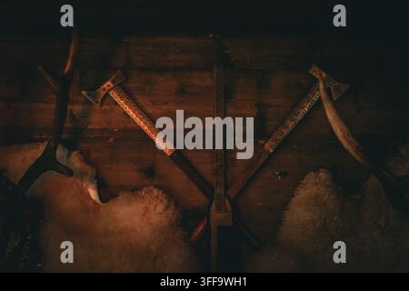Sammlung abgenutzter Äxte hängt an dunklem Holz neben weichen Fellen, die von einem schwachen Laternenlicht beleuchtet werden. Die Szene erinnert an Handwerkskunst, Waldarbeit und die raue Wildnis Stockfoto