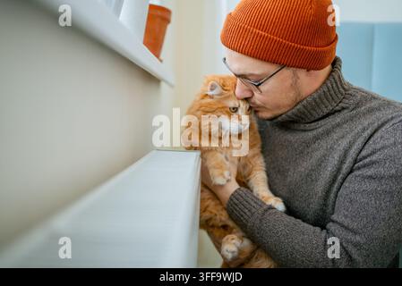 Mann mit orangefarbener Mütze und grauem Rollkragenpullover, der eine flauschige rote Katze am Kopf hält und küsst, zeigt Liebe und Zuneigung zwischen Haustierbesitzer und Haustier zu Hause Stockfoto