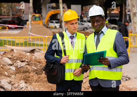 Ingenieur steht mit Baumeister, der den Bauprozess mit Hauszeichnungen auf der Struktur im Freien überwacht Stockfoto