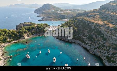 Drohnenpanorama über türkisfarbene Küstenbuchten in der Nähe von Rhodos, Griechenland, mit vor Anker liegenden Booten, felsigen Klippen, kleinen Stränden, bewaldeten Hügeln, einem Küstenparkplatz Stockfoto