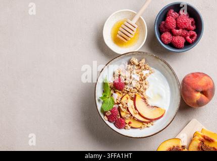 Flachlegung von griechischem Naturjoghurt mit Hafergranola oder Müsli, Pfirsich, Himbeere und Honig in einer Schüssel auf hellem Hintergrund mit frischen Früchten und Beeren. Stockfoto