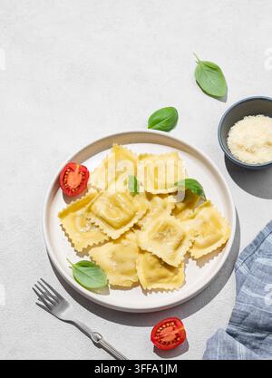Ravioli-Pasta mit Parmesan, Tomaten und Basilikum auf einem Teller vor einem hellen Hintergrund mit Schatten, Gabel und Käse. Traditionelles italienisches Hausgemachtes Essen con Stockfoto