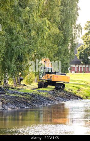 Schwerer gelber Bagger führt Umweltaufräumarbeiten am Flussufer und Kanalschonung an einem sonnigen Tag in einer grünen Parkumgebung durch. Stockfoto