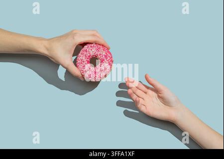 Hand hält einen glasierten rosa Donut zur anderen Hand auf blauem Hintergrund. Kreatives Konzept eines Mocks oder einer Werbung für ein schnelles Frühstück. Frontansicht und Kopie sp Stockfoto