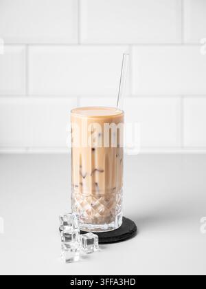 Kalter Latte-Kaffee in einem hohen Glas mit alternativer Milch und Strohhalm auf einem hellen Tisch vor einem weißen Ziegelhintergrund mit Eiswürfeln. Sommerlicher trendiger Erfrischungsgetränk Stockfoto