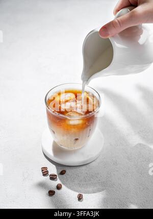 Kalter Latte-Kaffee in einem Glas, wobei eine Hand alternative Milch aus einem Milchkännchen über einen hellen Hintergrund mit Kaffeebohnen und Schatten gießt. Sommererfrischend Stockfoto