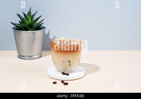 Kalter Latte-Kaffee in einem Glas mit Milch auf einem gelb-blauen Hintergrund mit Kaffeebohnen, Zimmerpflanze und Schatten. Sommerliches, trendiges, erfrischendes Getränk Stockfoto