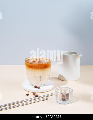 Kalter Latte-Kaffee in einem Glas mit Milch auf einem gelb-blauen Hintergrund mit Kaffeebohnen, Milchkanne und Schatten. Sommerliches, trendiges, erfrischendes Getränkekonzept. Stockfoto
