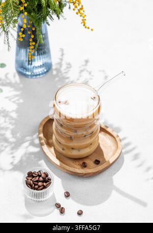 Kaltes Kaffeelatte oder Mokka in einem Glas mit Milch und Eis auf einem hellen Hintergrund mit einem Strauß Mimose, Kaffeebohnen und Morgenschatten. Das Konzept Stockfoto