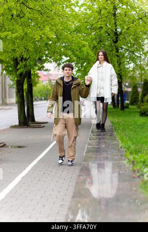 Junges Paar geht Hand in Hand entlang eines grünen Stadtrandsteins, reflektiert in Glas, und genießt ein romantisches Frühlingsdate im Freien. Stockfoto