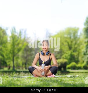 Junge Frau sitzt im Schneidersitz auf einer Yogamatte in einem Park und hält eine Wasserflasche Stockfoto