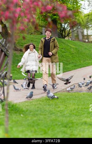 Ein glückliches junges Paar, das sich an den Händen hält und einen Parkweg entlangläuft, während im Frühling Tauben um sie herumfliegen. Stockfoto