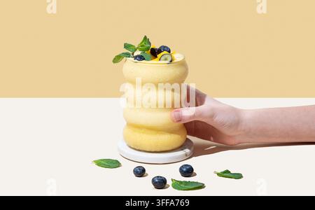 Hand, die ein Glas Smoothie mit Mango, Blaubeere und Minze in einem Glas auf einem gelb-weißen Hintergrund hält. Konzept für gesunde Ernährung und Detox-Diät. Fro Stockfoto