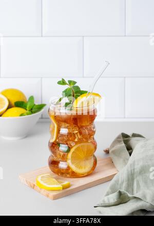 Eistee mit Zitrone, Eis und Minze in einem Glas auf der Küchenarbeitsplatte mit Zitrusfrüchten und weißem Ziegelwandhintergrund. Trendiges Konzept der Erfrischung Stockfoto