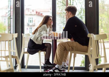 Junges Paar genießt Kaffee und Gespräch an einem hellen Cafétisch an großen Fenstern und teilt einen entspannten Moment zusammen. Stockfoto