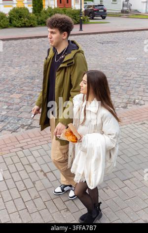 Junges Paar geht Hand in Hand auf Kopfsteinpflasterstraße, Frau hält frische Backwaren in einer Papiertüte während eines lockeren Stadt-Dates. Stockfoto