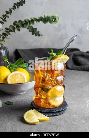 Eistee mit Zitrone, Eis und Minze in einem Glas auf grauem Hintergrund mit Zitrusfrüchten und Serviette. Trendiges Konzept für ein erfrischendes Getränk oder Limonade. Fr Stockfoto