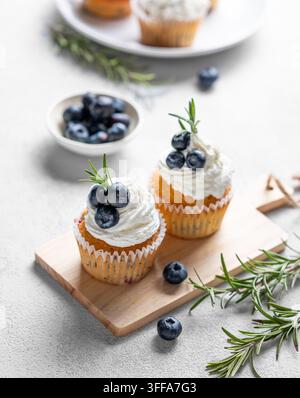 Cupcakes mit Frischkäseglasur, frischen Blaubeeren und Rosmarin auf einem Holzbrett auf einem hellen Hintergrund mit frischen Beeren. Konzept eines köstlichen Stockfoto