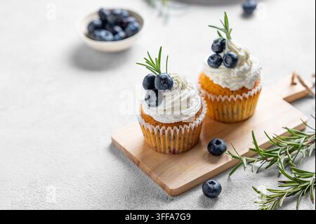 Cupcakes mit Frischkäseglasur, frischen Blaubeeren und Rosmarin auf einem Holzbrett auf einem hellen Hintergrund mit frischen Beeren. Konzept eines köstlichen Stockfoto