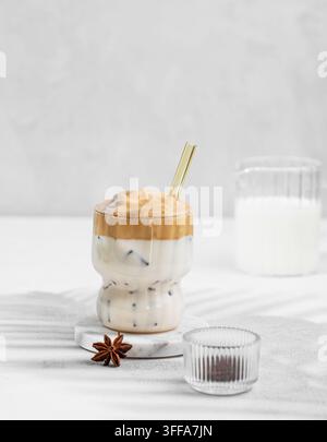 Dalgona-Kaffee in einem Glas auf hellem Hintergrund mit Milchkanne und Gewürzen. Koreanisches geschlagenes Getränk mit Instantkaffee, Zucker, Milch und Eis. Süß und trendig Stockfoto