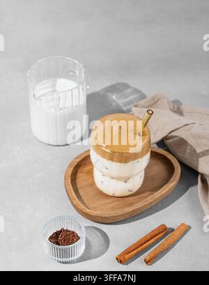 Koreanisches geschlagenes Getränk Dalgona-Kaffee auf einem Holzbrett auf einem Betonhintergrund mit Gewürz- und Milchkanne. Getränk mit Instantkaffee, Zucker, Milch und Eis Stockfoto