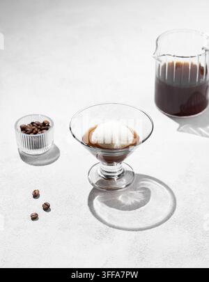 Affogato-Kaffee mit einer Kugel Vanilleeis und Espresso in einem Glas auf hellem Hintergrund mit Kaffeebohnen, Kanne und Schatten. Klassisches italienisches Des Stockfoto
